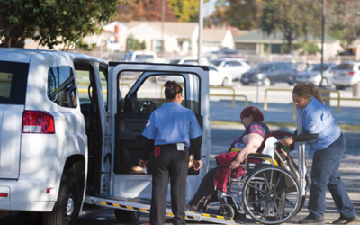 Woman in a non-motorized wheelchair is being assisted by two uniformed individuals into an accessible van with ramp.